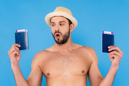 Excited shirtless man in straw hat holding passports and boarding passes isolated on blueの写真素材