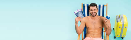 Smiling shirtless man holding passports and canned drink on deck chair near suitcase on blue background, bannerの写真素材
