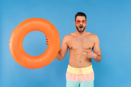 Astonished man in sunglasses pointing at inflatable ring isolated on blueの写真素材