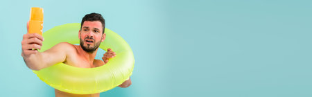 Man with sunburn holding inflatable ring and sunscreen isolated on blue, banner