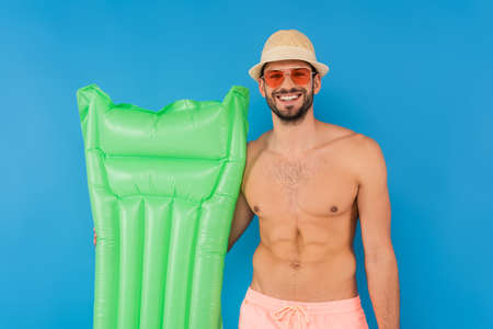 Shirtless man in sun hat and sunglasses holding inflatable mattress isolated on blueの写真素材