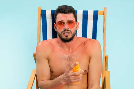 Worried shirtless man with sunburn holding sunscreen on deck chair isolated on blue