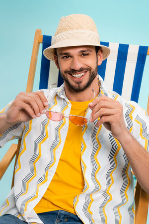 Positive man in straw hat holding sunglasses on deck chair isolated on blue