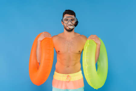 Shirtless man in swimming goggles holding inflatable rings isolated on blueの写真素材