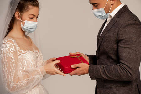 man in medical mask presenting gift box to young bride isolated on grayの写真素材