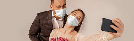 bride in medical mask taking selfie with groom isolated on gray, bannerの写真素材