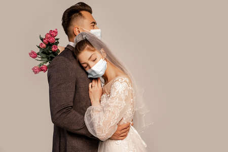 man in medical mask embracing bride leaning on him with closed eyes isolated on gray