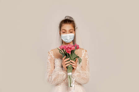 bride in medical mask holding bouquet of fresh tulips isolated on grayの写真素材