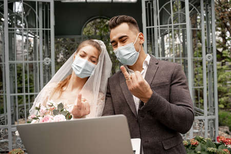 newlyweds in protective masks showing wedding rings during video chat on laptopの写真素材