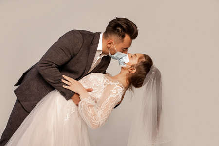 man in medical masks kissing elegant bride isolated on gray with lilac shade