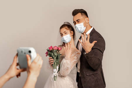 newlyweds in medical masks showing wedding rings near blurred photographer isolated on grayの写真素材