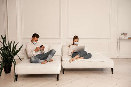 young couple in protective masks using gadgets on sofa in living roomの写真素材