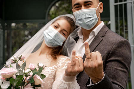couple of newlyweds in safety masks showing wedding rings at cameraの写真素材