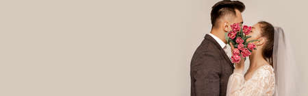 young newlyweds kissing while obscuring faces with tulips isolated on gray, bannerの写真素材