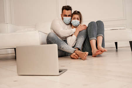 scared couple in medical masks watching horror film on laptop on floorの写真素材
