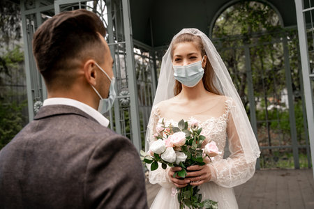 young bride in medical mask holding wedding bouquet near groom on blurred foregroundの写真素材