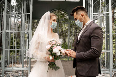 young man in medical mask putting wedding ring on finger of brideの写真素材