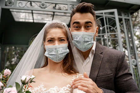 excited newlyweds in safety masks with bride and groom lettering looking at camera in parkの写真素材