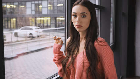 serious young adult woman standing near window in modern loftの写真素材