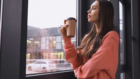 young adult woman holding paper cup and looking at window in modern loftの写真素材