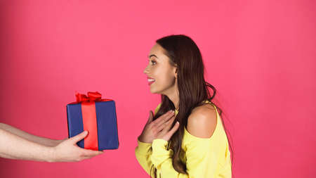 amazed young adult woman standing near male hands with gift box isolated on pinkの写真素材