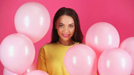 smiling young adult woman in yellow blouse among balloons isolated on pinkの写真素材
