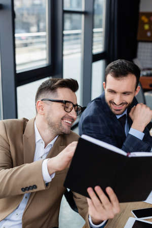 Smiling businessman holding blurred notebook near colleague