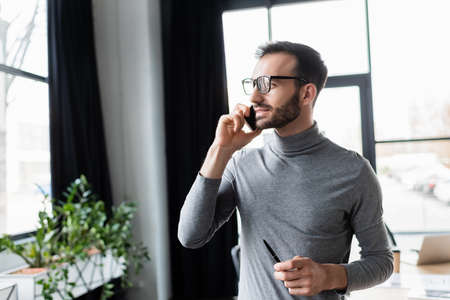 Businessman holding pen while talking on smartphone in officeの写真素材