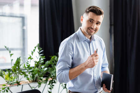 Cheerful businessman with pen and notebook looking at camera in officeの写真素材
