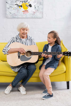 Cheerful grandmother and kid playing acoustic guitar on couchの写真素材