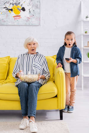 Shocked kid holding remote controller near grandmother with popcornの写真素材