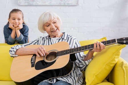 Elderly woman smiling while playing acoustic guitar near blurred kid on couchの写真素材