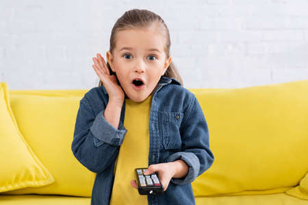 Astonished kid with remote controller sitting on couchの写真素材