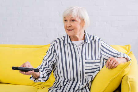 Cheerful senior woman with remote controller watching tv at homeの写真素材