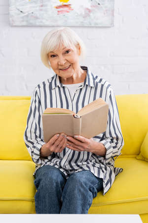 Senior woman with book smiling at camera on couch
