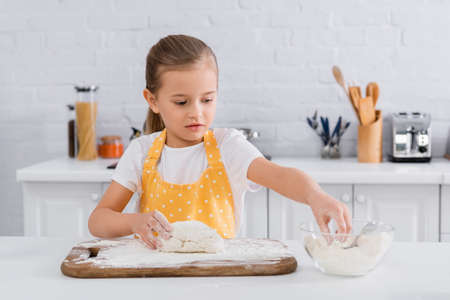 Kid in apron taking flour near dough on cutting boardの写真素材