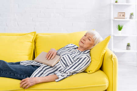 Elderly woman holding book while sleeping on couchの写真素材