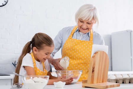 Positive kid and grandmother pouring milk in bowl near digital tablet and flourの写真素材