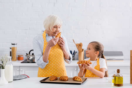 Senior granny smelling croissant near granddaughter in kitchenの写真素材