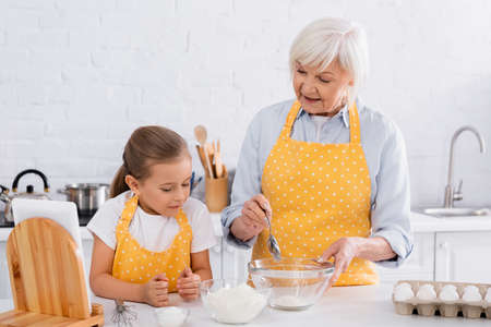 Smiling grandmother holding spoon near flour, eggs and granddaughter in kitchenの写真素材
