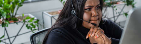 pensive african american plus size operator in headset with microphone looking at computer monitor,...
