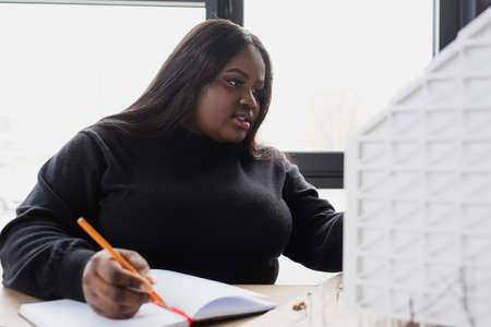 african american plus size designer writing in notebook while looking at house modelの写真素材