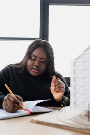 african american plus size designer writing in notebook near blurred house model