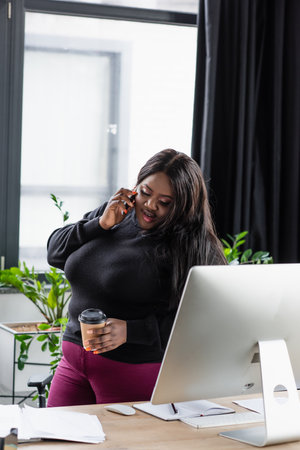 cheerful african american plus size businesswoman talking on smartphone and holding coffee to go in officeの写真素材