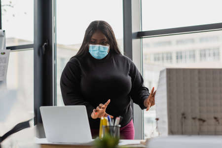 african american plus size businesswoman in medical mask gesturing during video call on laptop in officeの写真素材
