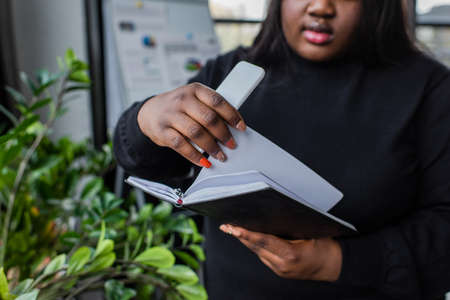 cropped view of african american plus size woman holding notebook and smartphone in officeの写真素材