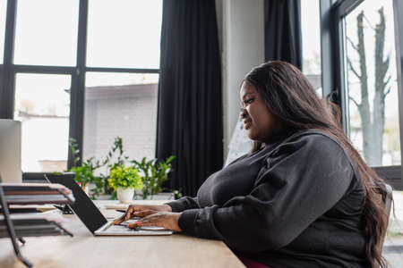 happy african american plus size woman typing on laptop keyboard in officeの写真素材