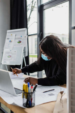 african american plus size engineer in medical mask looking at blueprint near laptop and house model in officeの写真素材