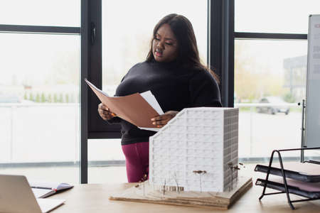 brunette african american plus size engineer holding folder near house model on desk