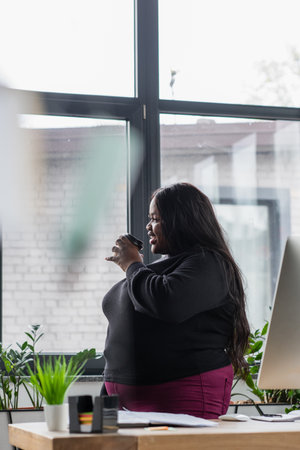 smiling african american plus size businesswoman holding coffee to go in officeの写真素材